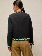 A woman shown from the back wearing a dark charcoal knit cardigan with green and white chevron trim, paired with tan pants, against a neutral beige background.