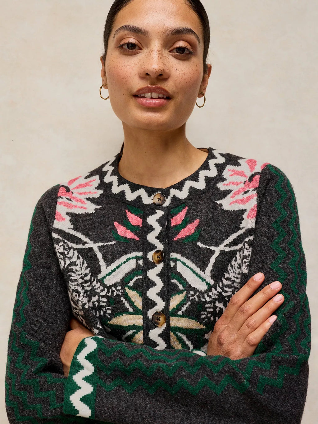 A woman wearing a dark gray knit cardigan with a colorful Fair Isle pattern featuring pink, green, white, and beige motifs, posed with arms crossed against a neutral beige background.