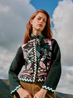 A woman outdoors wearing a dark green patterned knit cardigan with a matching neck scarf, brown corduroy trousers, and gold hoop earrings, set against a cloudy blue sky.