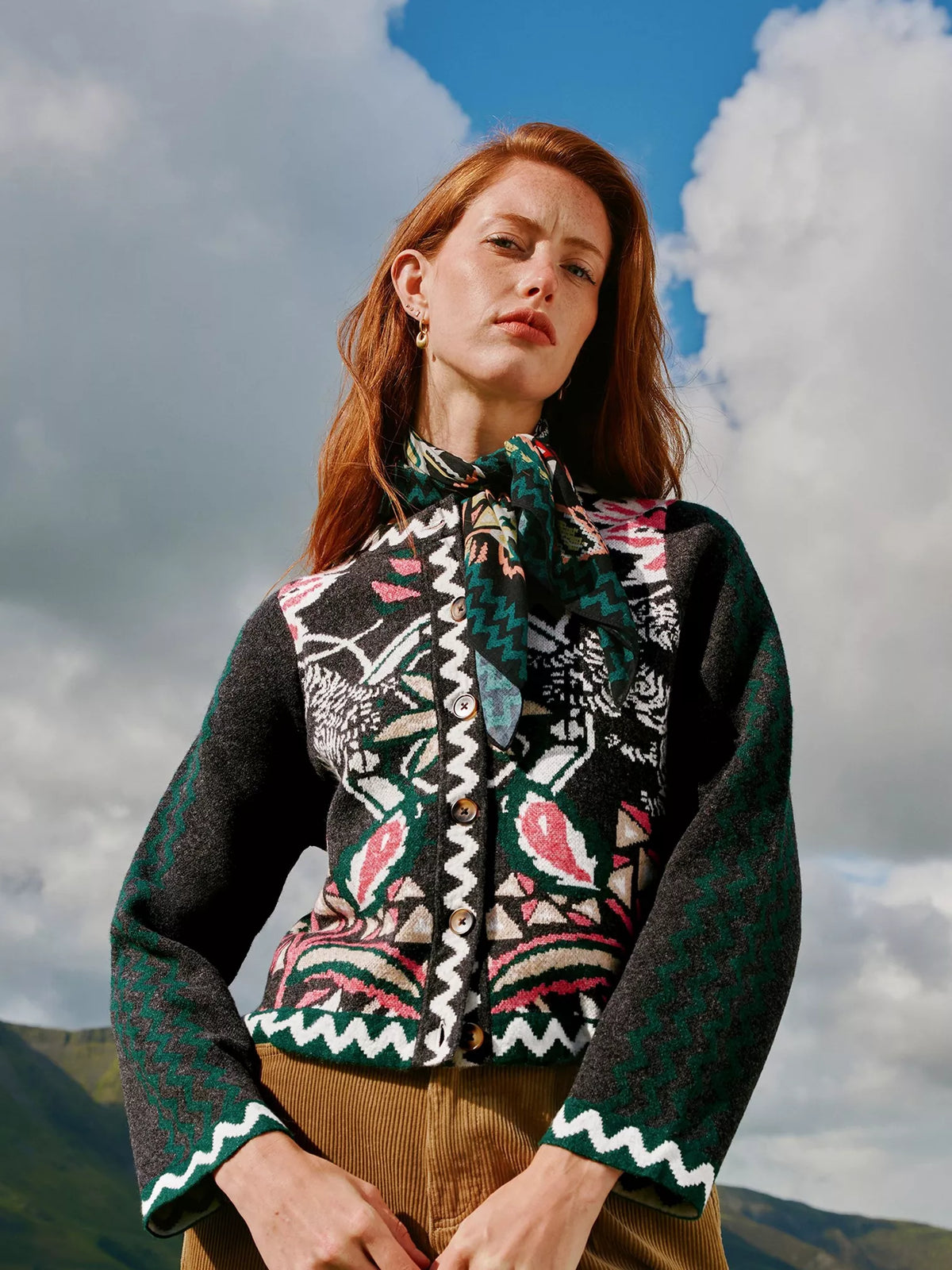 A woman outdoors wearing a dark green patterned knit cardigan with a matching neck scarf, brown corduroy trousers, and gold hoop earrings, set against a cloudy blue sky.