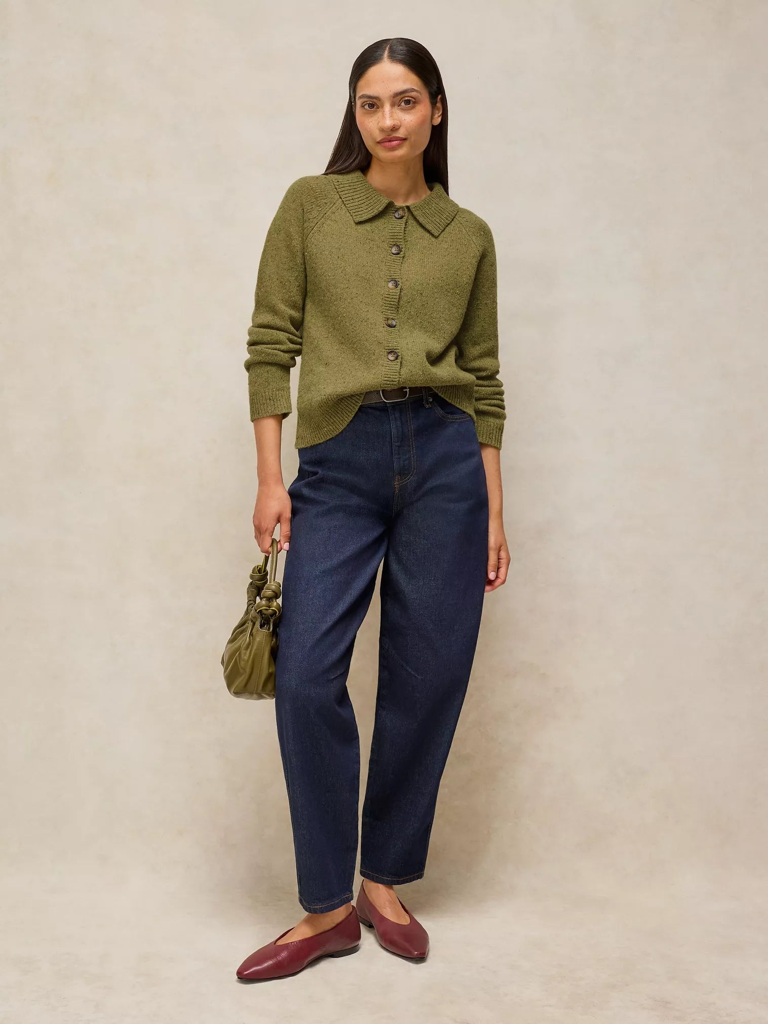A woman stands against a warm beige textured backdrop wearing Shepherd Collar Cardi – Mid Chartreuse, dark blue jeans, burgundy flats, and holding a small olive green handbag.