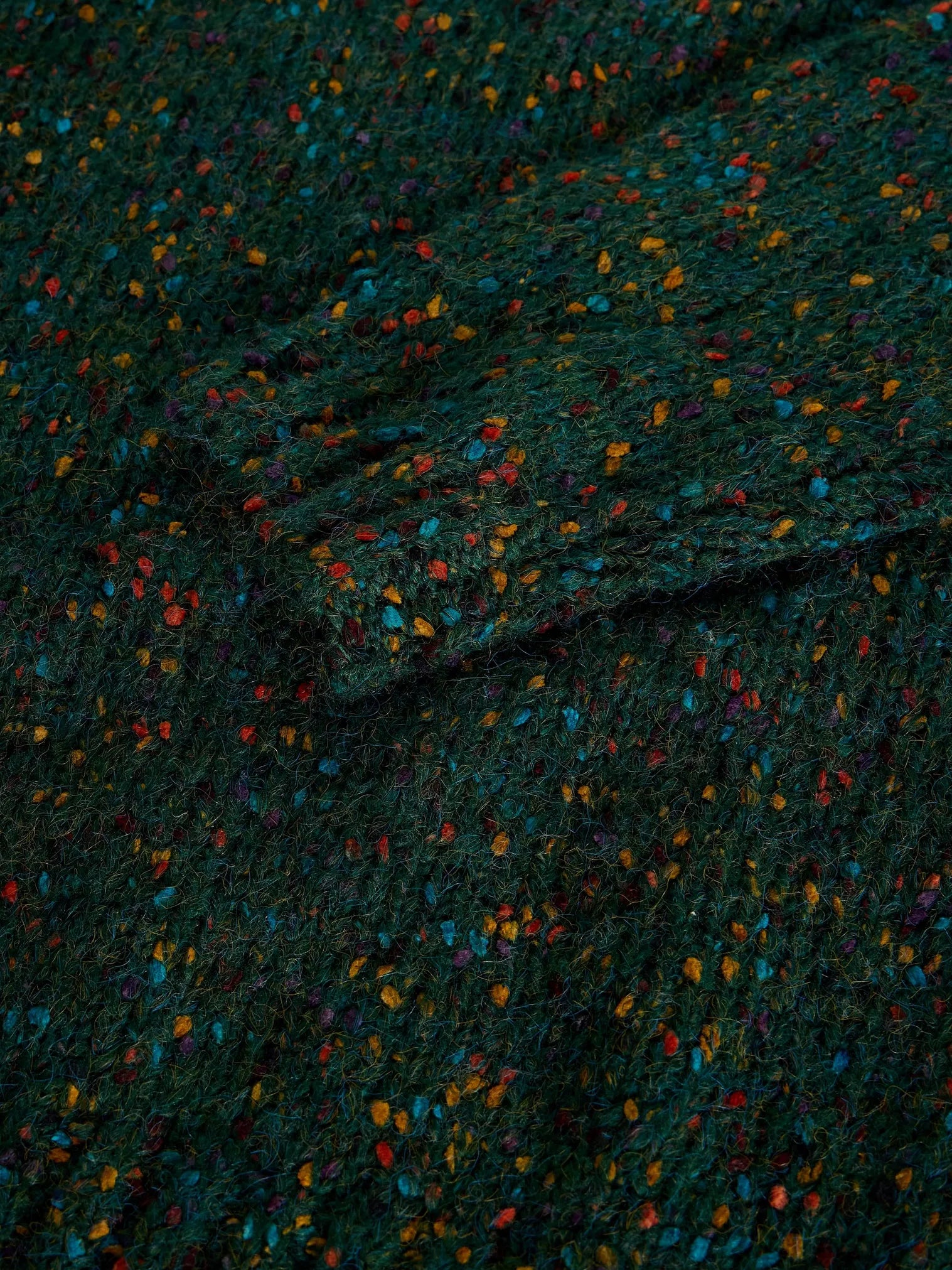 A close-up of a dark green textured fabric with a multicolor speckled pattern of small orange, blue, red, and yellow flecks.