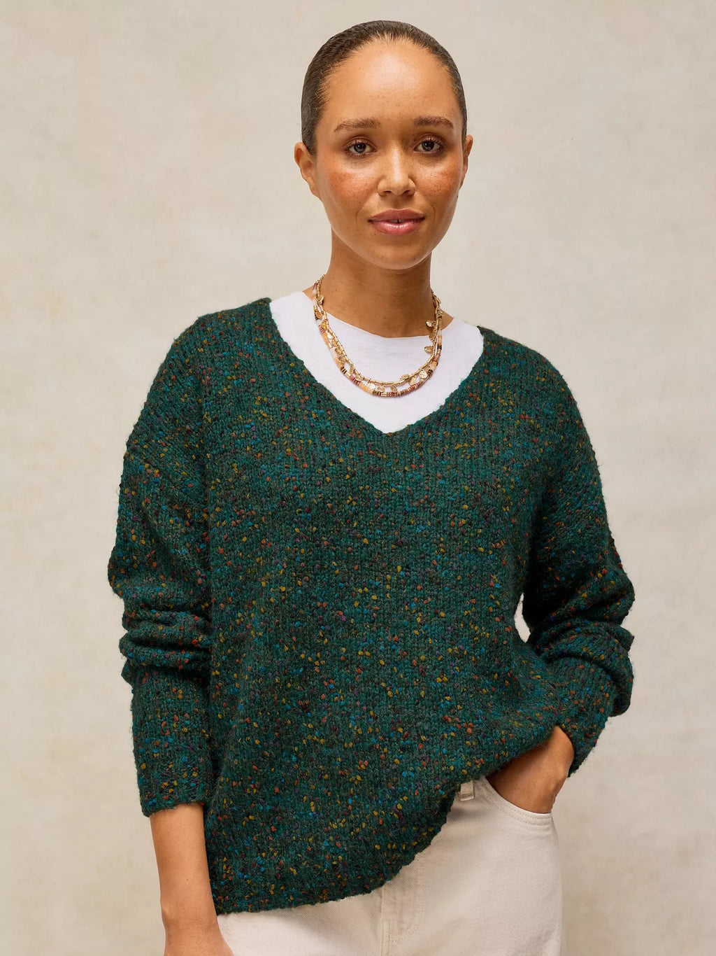 A woman wearing a dark green marled knit sweater with a white V-neck undershirt and cream pants, standing against a neutral textured beige background.