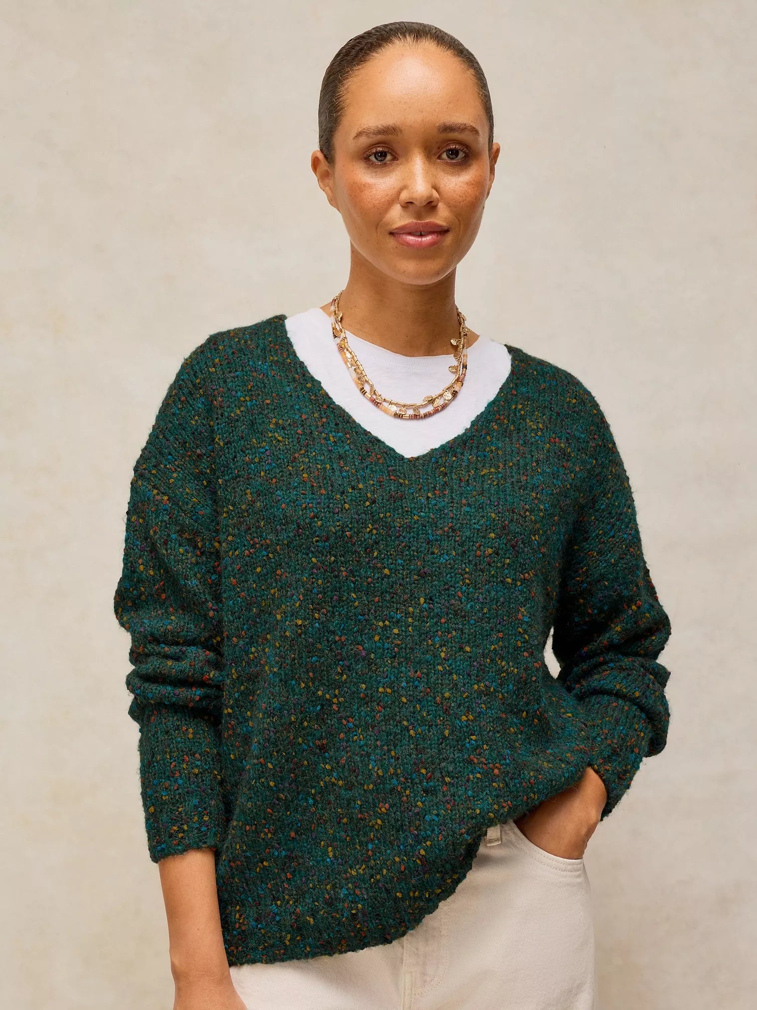 A woman wearing a dark green marled knit sweater with a white V-neck undershirt and cream pants, standing against a neutral textured beige background.