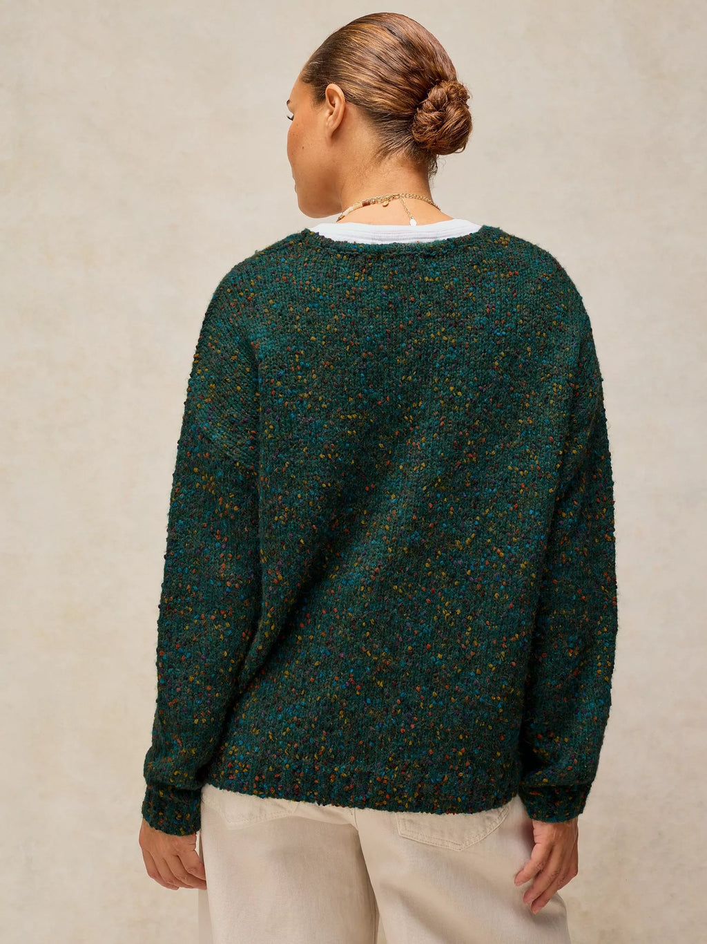 A woman viewed from the back wearing a dark green textured knit sweater with subtle multicolor flecks and light beige pants, set against a neutral beige background.