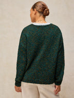 A woman viewed from the back wearing a dark green textured knit sweater with subtle multicolor flecks and light beige pants, set against a neutral beige background.