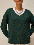 A person wearing a dark green marled knit sweater over a white crewneck shirt, accessorized with a chunky gold chain necklace, against a neutral beige background.