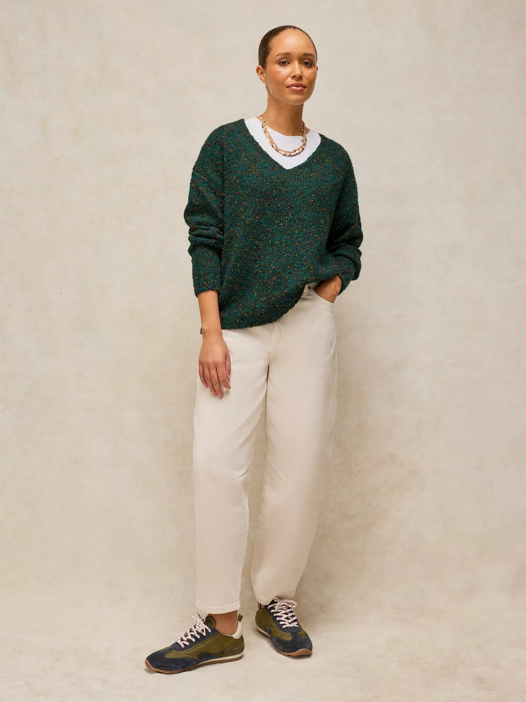 A woman modeling a dark green textured knit V-neck sweater with a white collar, paired with cream trousers and dark blue sneakers, standing against a textured beige backdrop.
