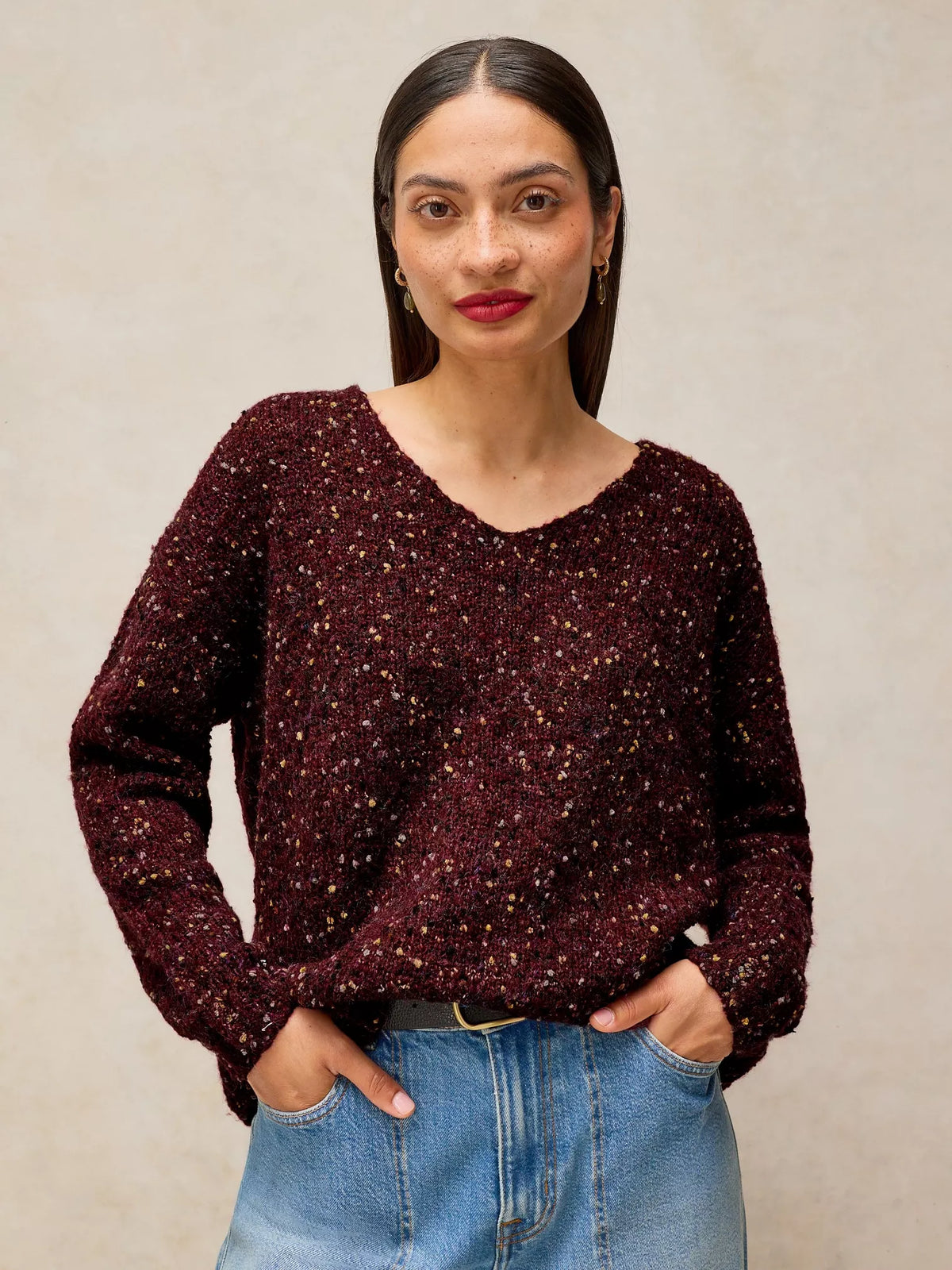 A woman wearing a dark burgundy long-sleeve knit sweater with a textured, sparkly glitter pattern, paired with light blue jeans, standing against a neutral beige background.