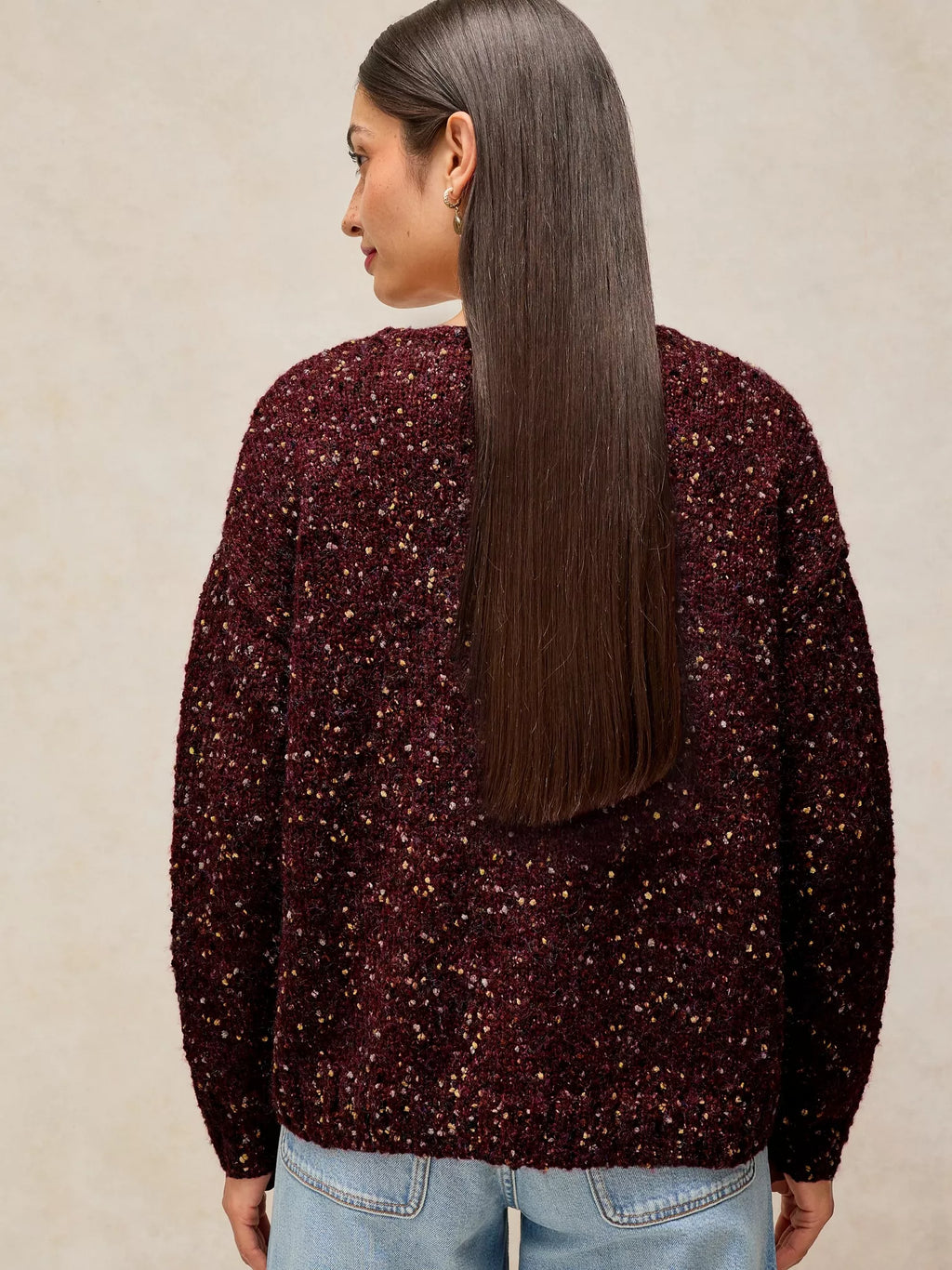 A woman shown from the back wearing a dark burgundy fuzzy knit sweater covered in small gold sequins, paired with light blue jeans, against a neutral beige background.