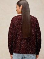 A woman shown from the back wearing a dark burgundy fuzzy knit sweater covered in small gold sequins, paired with light blue jeans, against a neutral beige background.
