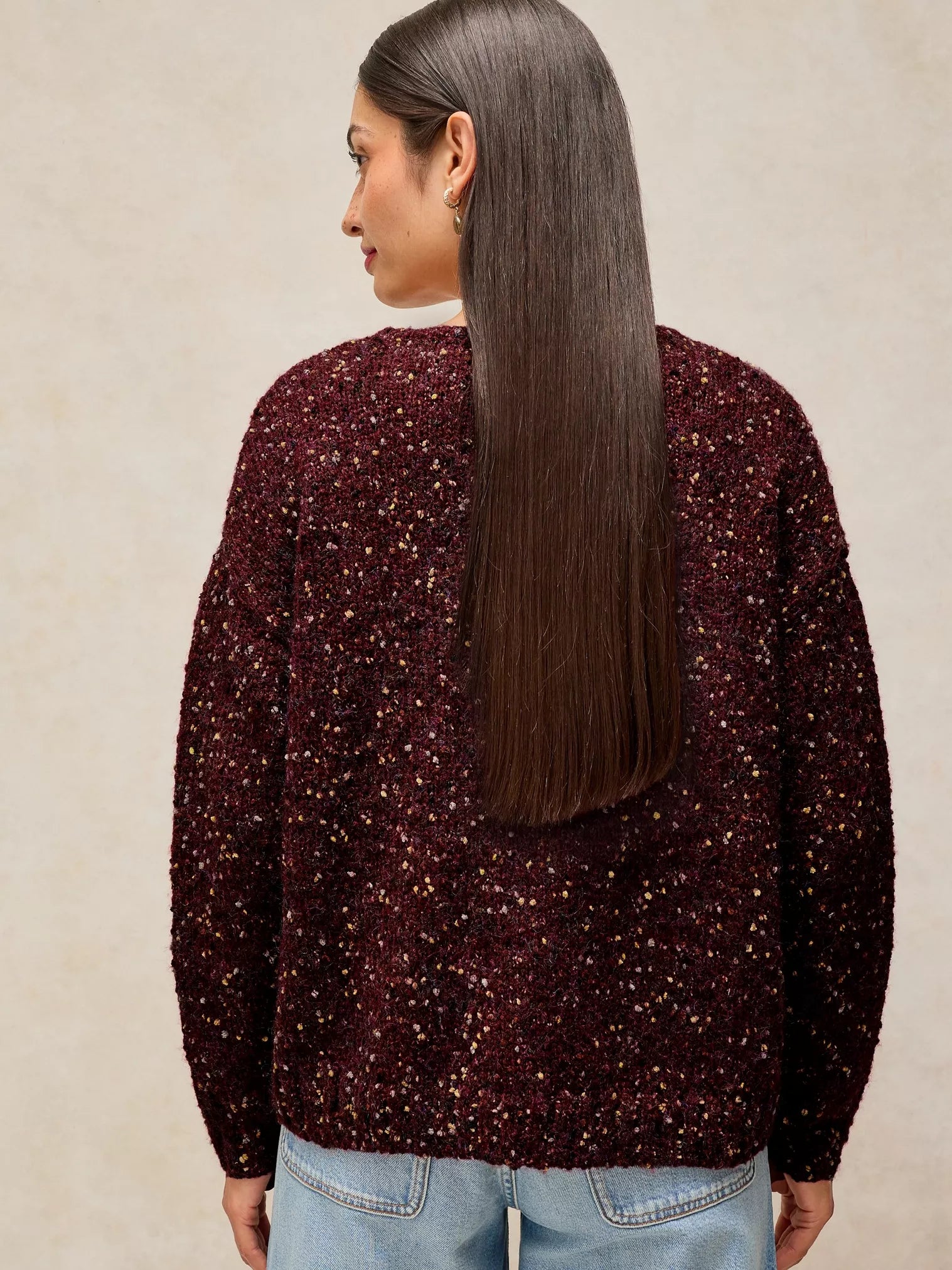 A woman shown from the back wearing a dark burgundy fuzzy knit sweater covered in small gold sequins, paired with light blue jeans, against a neutral beige background.
