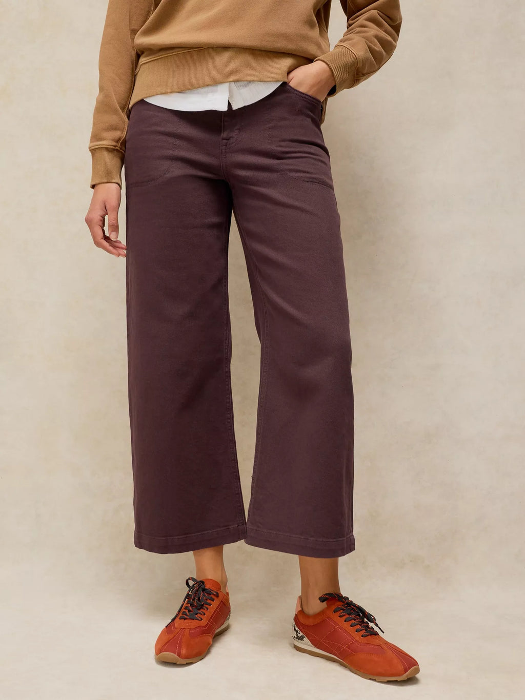 A person wearing dTIA brown WIDE LEG CROP JEAN , styled with a tan sweatshirt, white shirt, and rust-colored sneakers, standing against a textured beige studio backdrop.