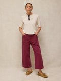 A model stands against a textured beige studio backdrop wearing a cream short-sleeve knit top, wide-leg cropped trousers in a deep burgundy, and tan lace-up shoes. Sunglasses hang from the neckline.