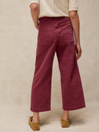 A person shown from the back wearing high-waisted, wide-leg cropped pants in a deep burgundy or maroon shade, paired with a cream short-sleeve top and tan wedge sandals, against a textured beige studio backdrop.