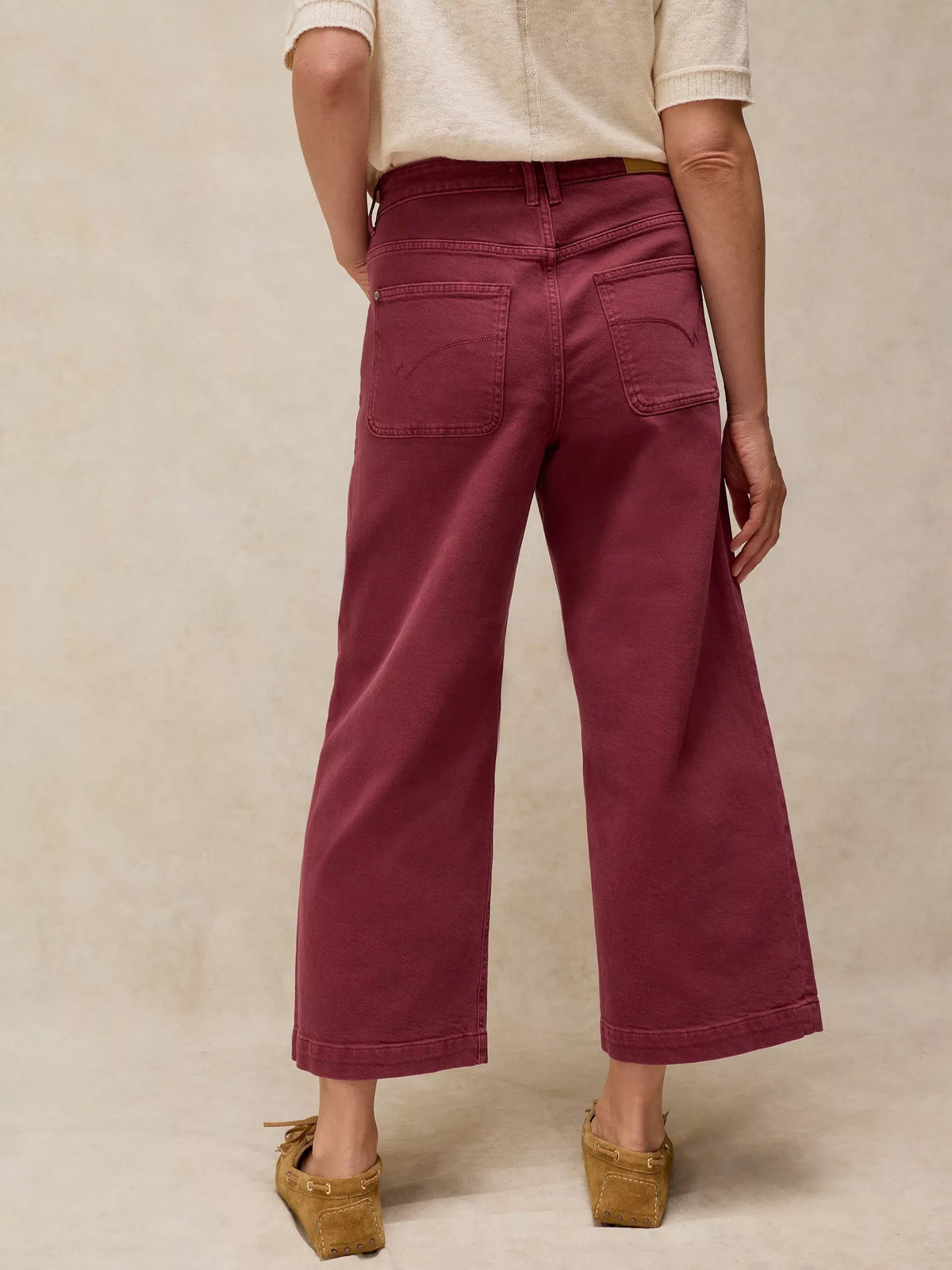 A person shown from the back wearing high-waisted, wide-leg cropped pants in a deep burgundy or maroon shade, paired with a cream short-sleeve top and tan wedge sandals, against a textured beige studio backdrop.
