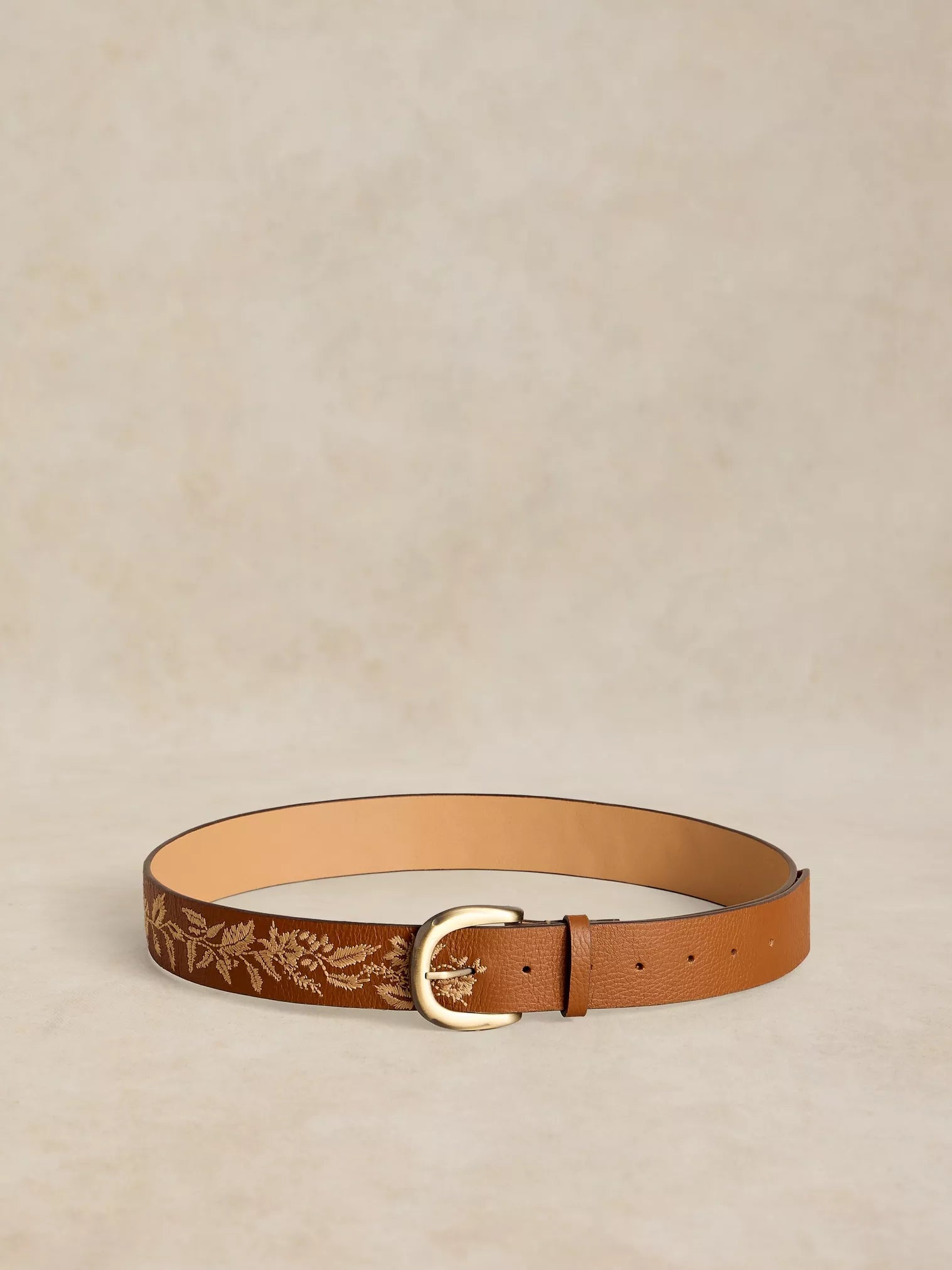 A tan leather belt with a gold-toned buckle and a debossed floral pattern, displayed on a textured beige background.