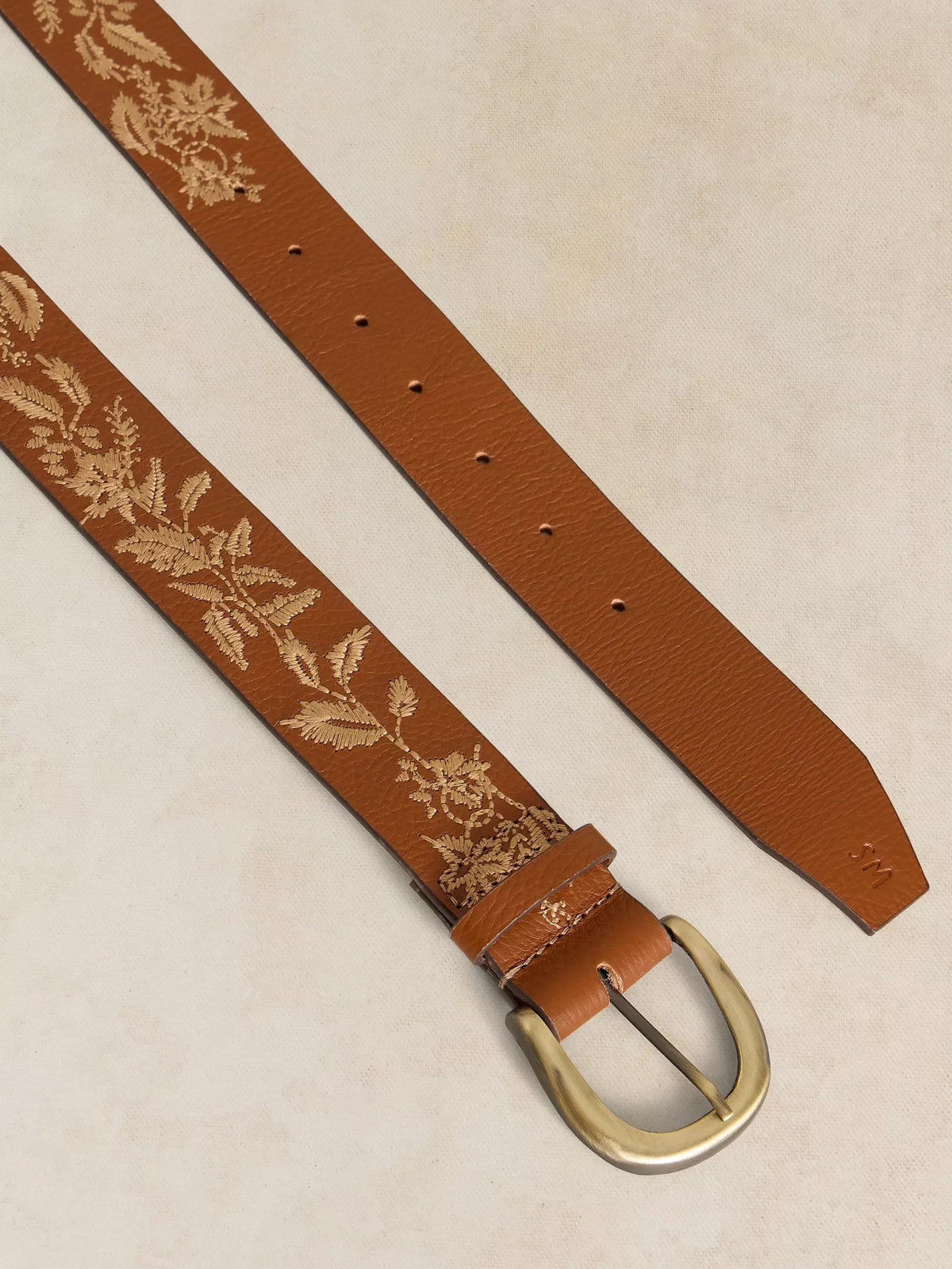 A brown leather belt with an embroidered beige floral vine pattern and a polished gold-tone buckle, laid diagonally on a textured beige background.