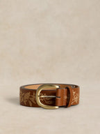 A brown leather belt with a gold floral pattern and a gold-tone buckle, displayed on a textured beige background.