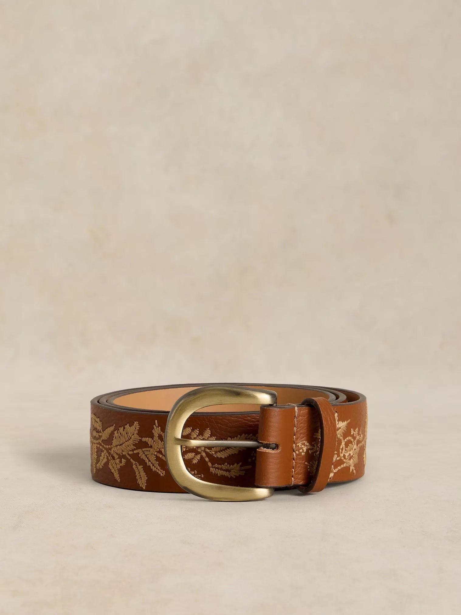 A brown leather belt with a gold floral pattern and a gold-tone buckle, displayed on a textured beige background.