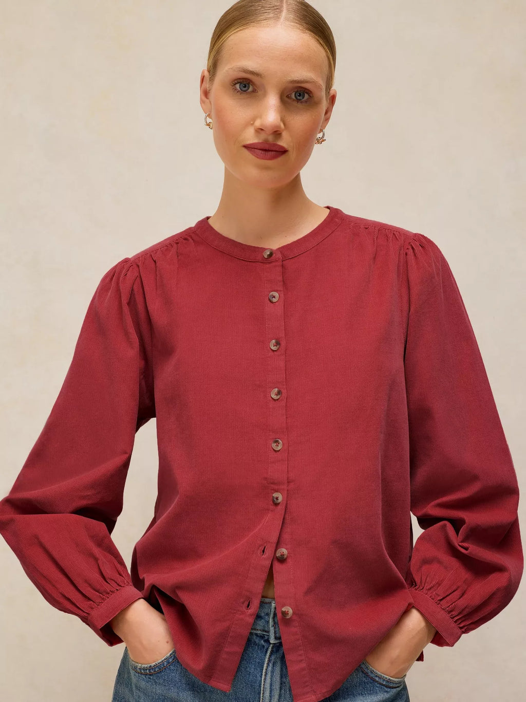 A woman wearing a long-sleeve button-front LANA dusty PINk BABY CORD TOP , paired with blue jeans, against a neutral beige background.