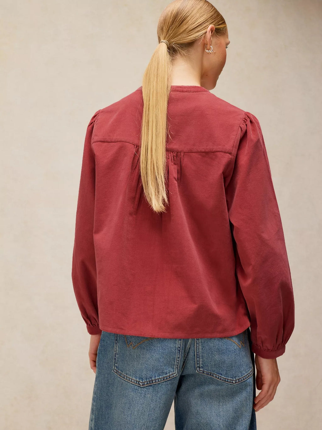 A woman viewed from the back wearing a LANA dusty PINk BABY CORD TOP with gathered shoulders and blue jeans, standing against a neutral beige textured wall.