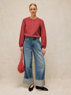 A woman stands against a textured beige studio backdrop wearing a LANA dusty PINk BABY CORD TOP  with front buttons, wide-leg blue jeans with rolled cuffs, black flats, and holding a red handbag.
