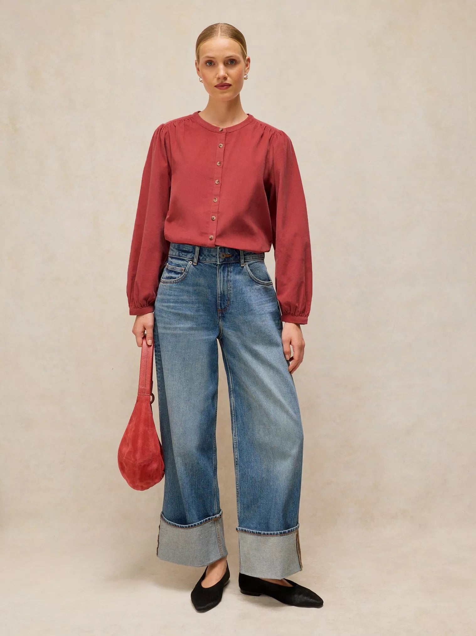 A woman stands against a textured beige studio backdrop wearing a LANA dusty PINk BABY CORD TOP  with front buttons, wide-leg blue jeans with rolled cuffs, black flats, and holding a red handbag.