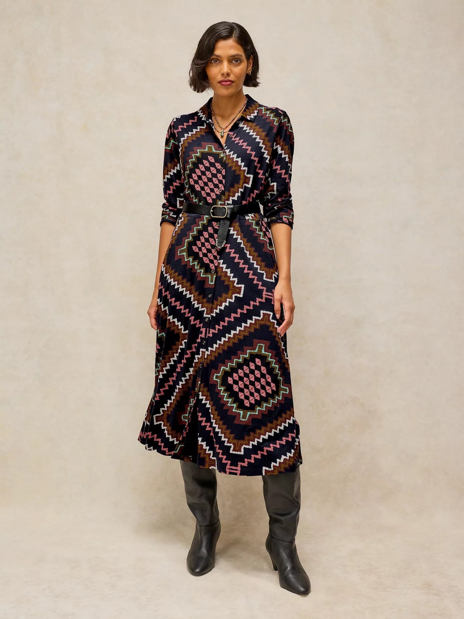 A woman modeling Rua Jersey Dress in Navy Multi with a geometric pattern in black, pink, brown, and white, featuring a matching black belt, styled with black boots against a textured beige background.