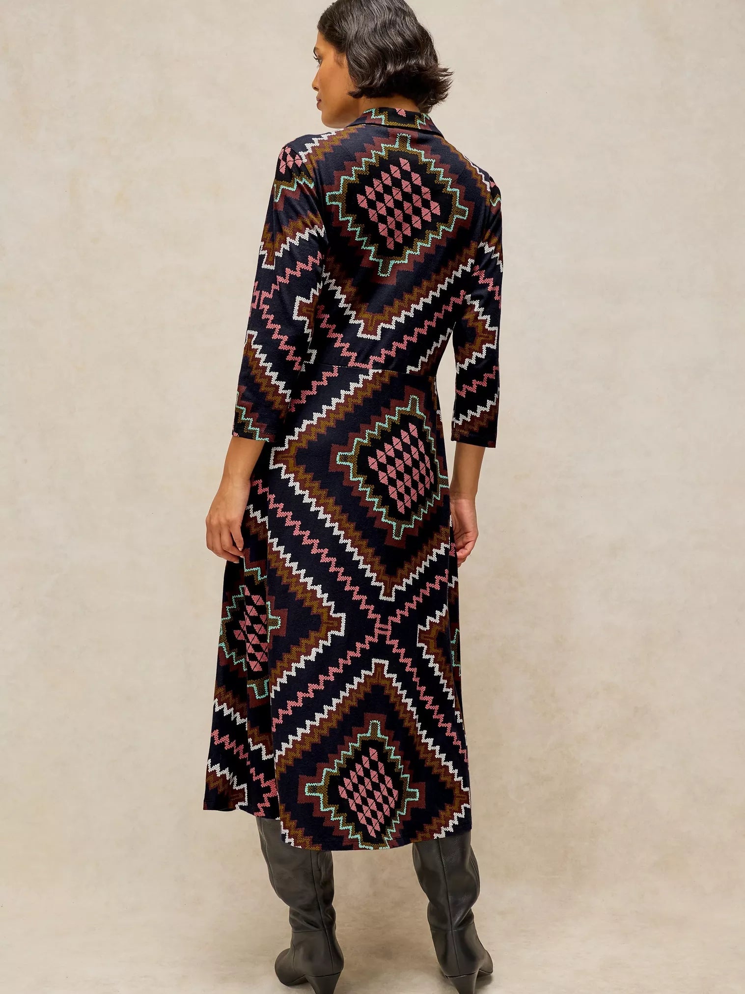 A woman shown from the back Rua Jersey Dress in Navy Multi with a geometric Southwestern-style print in black, pink, white, and brown, paired with dark gray boots, against a neutral beige background.
