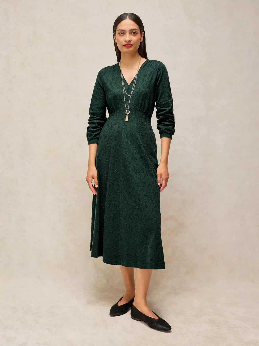 A woman modeling a dark green long-sleeve midi dress with a V-neckline, standing against a neutral textured beige backdrop.