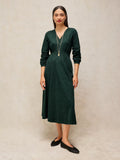 A woman modeling a dark green long-sleeve midi dress with a V-neckline, standing against a neutral textured beige backdrop.