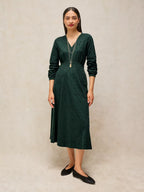 A woman modeling a dark green long-sleeve midi dress with a V-neckline, standing against a neutral textured beige backdrop.