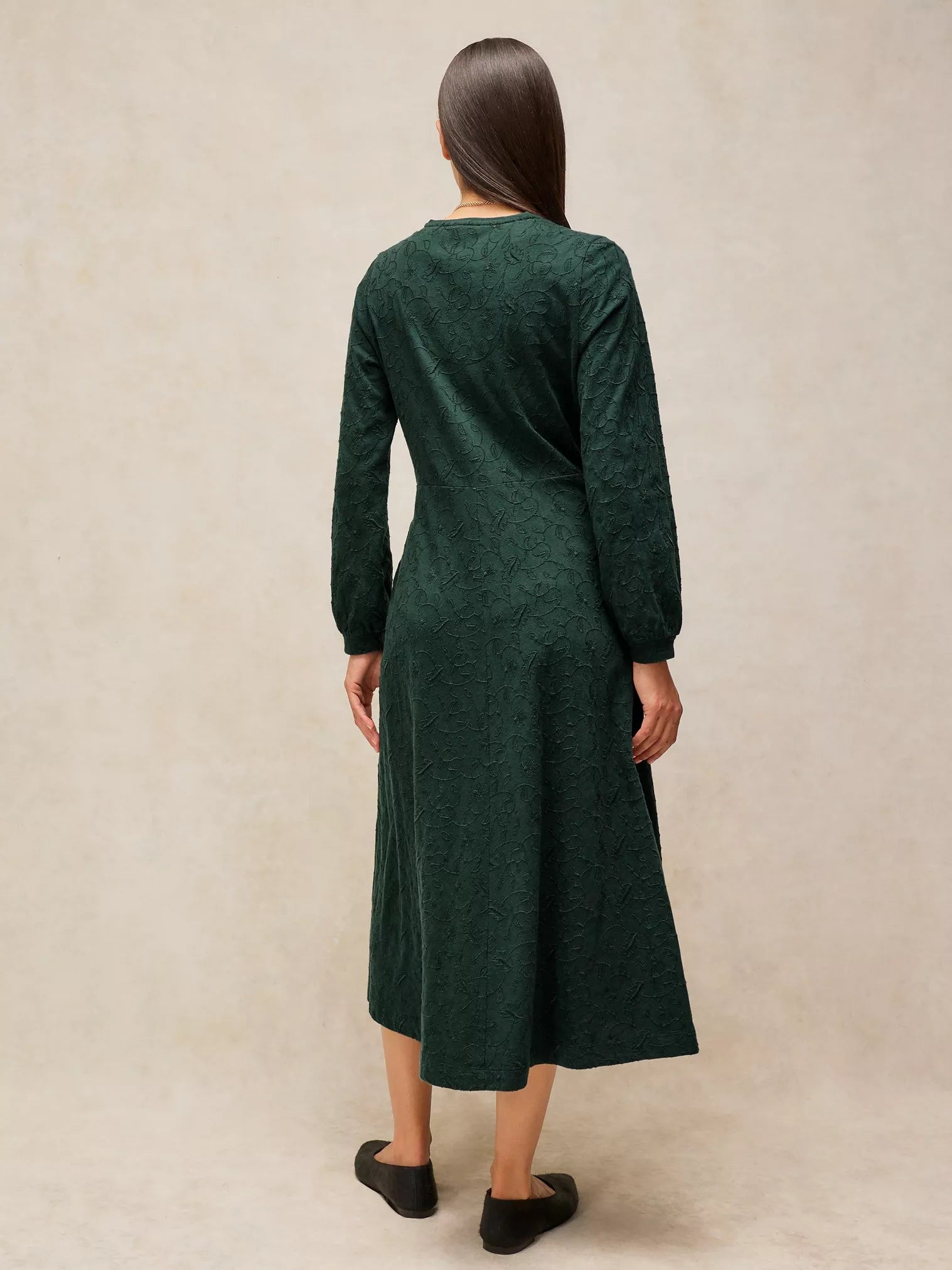 A woman viewed from the back wearing a dark green long-sleeve midi dress with a textured knit pattern, standing against a neutral beige studio backdrop.