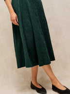 A dark green midi skirt with a textured floral lace pattern, shown on a model wearing black flats against a neutral beige background.