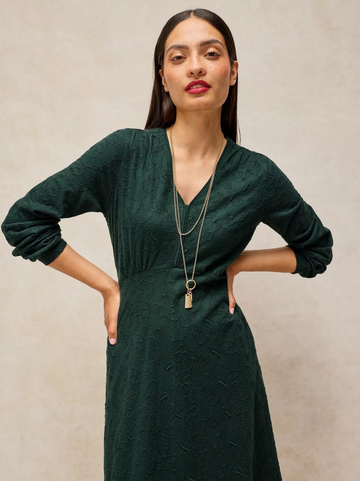 A woman modeling a dark forest green textured long-sleeve dress with a V-neckline, paired with a double-layered gold pendant necklace, set against a neutral beige textured wall.