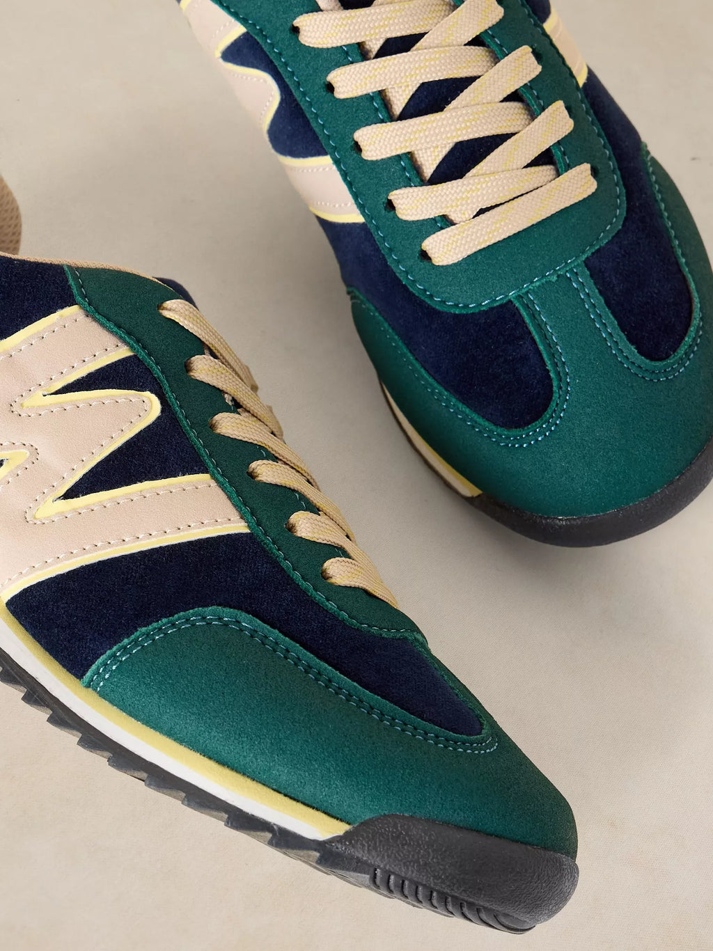 A close-up of a pair of A Lucy Trainer sneaker in Blue multi in green suede and navy blue mesh with cream laces and side stripes, set against a light beige background.