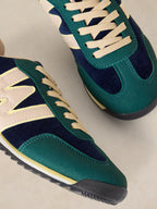 A close-up of a pair of A Lucy Trainer sneaker in Blue multi in green suede and navy blue mesh with cream laces and side stripes, set against a light beige background.