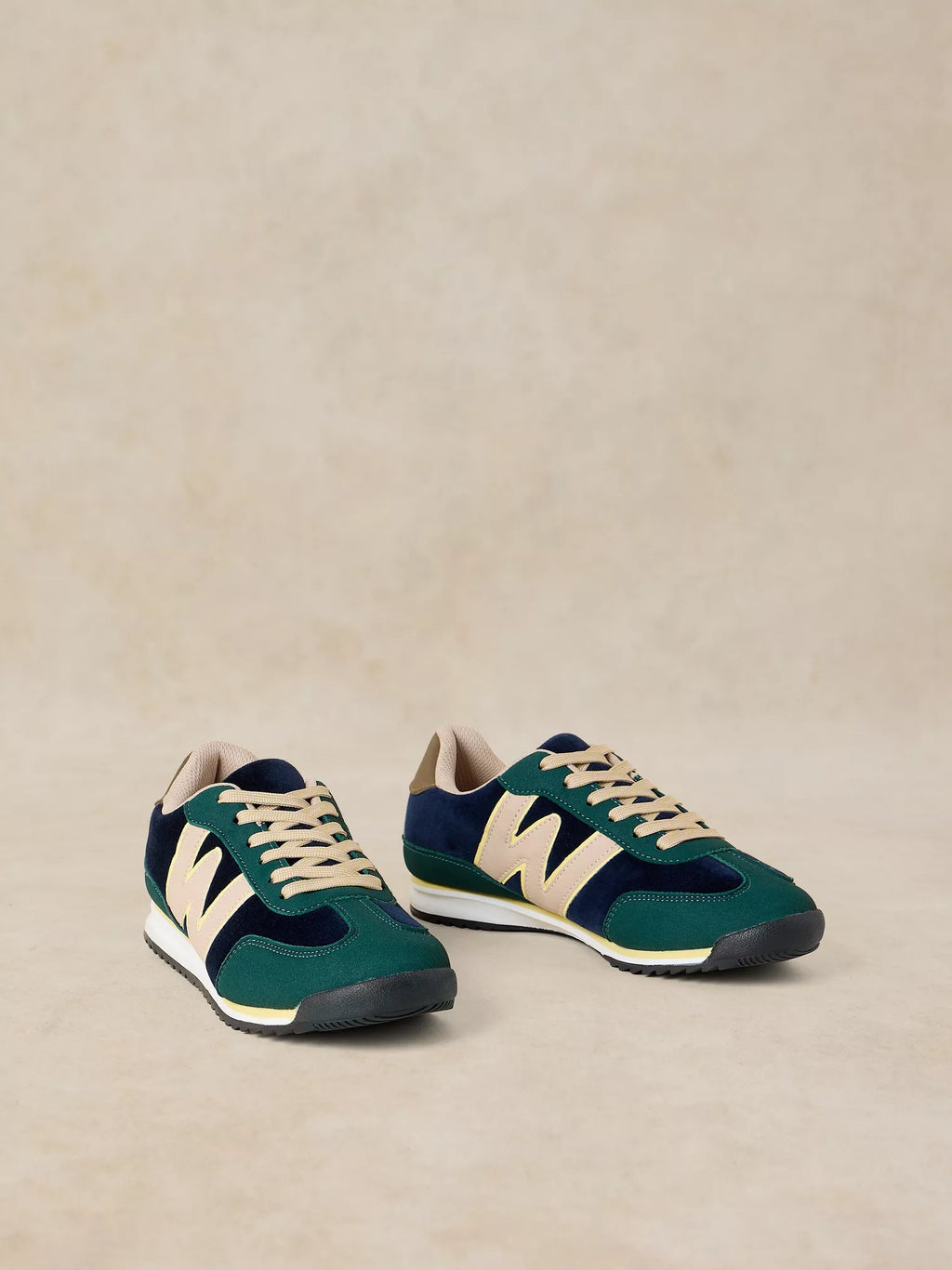 A pair of A Lucy Trainer sneaker in Blue multi  in dark green suede and navy blue mesh, featuring beige leather 'N' logos and beige laces, set against a neutral beige background.
