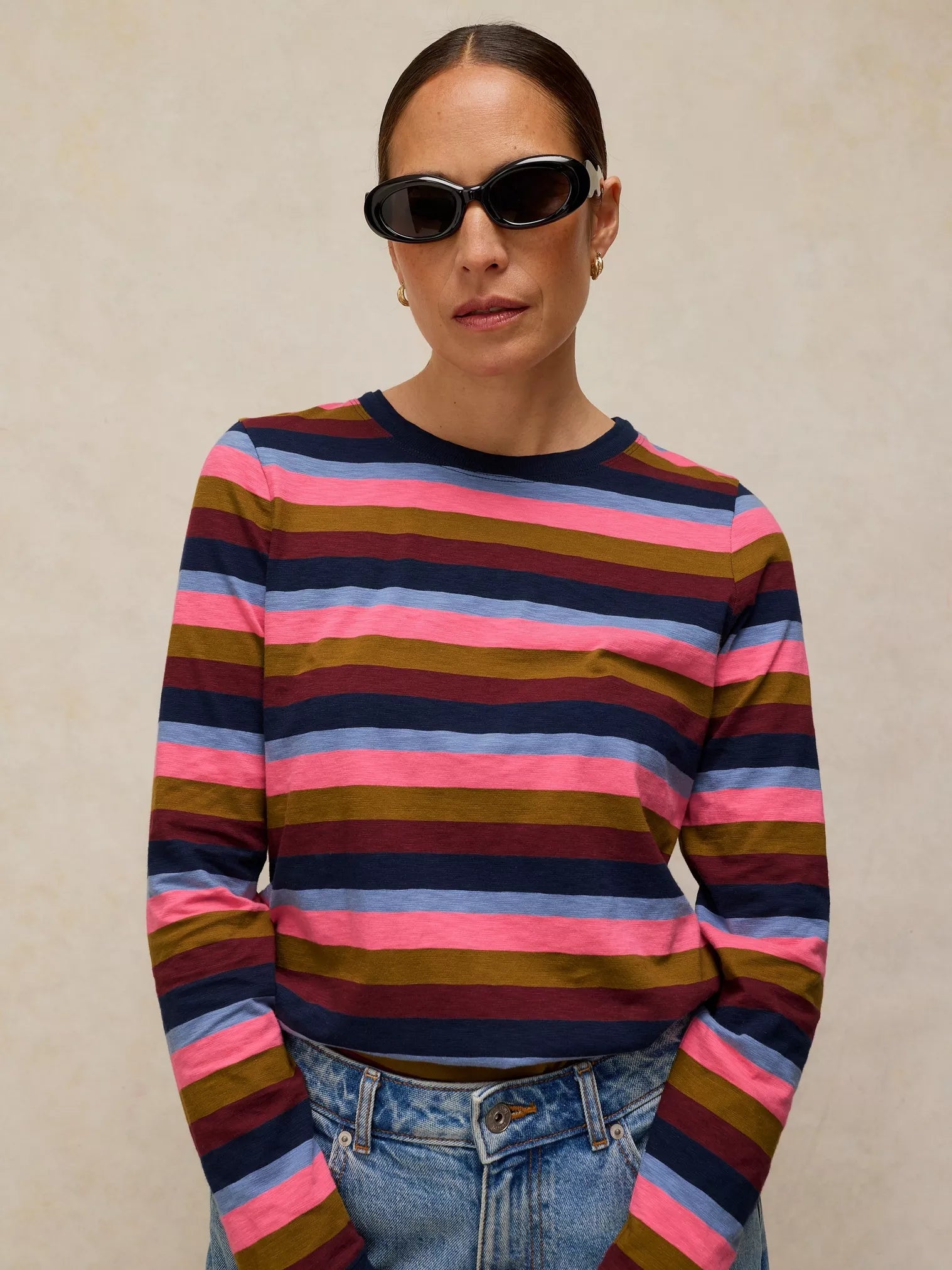 A woman wearing a long-sleeve CLARA STRIPE LS TEE
in PINK MULTI paired with blue jeans and black sunglasses, against a textured beige background.