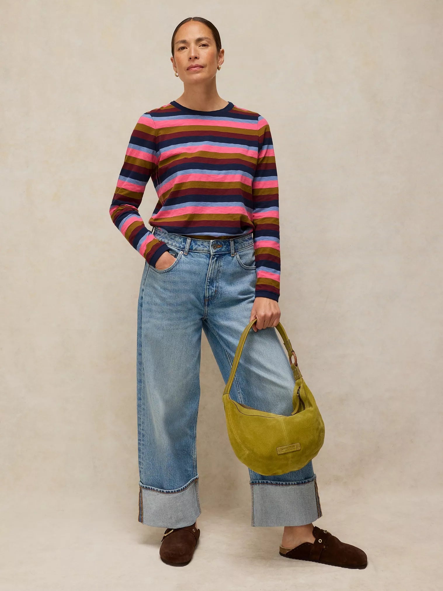 Person wearing a CLARA STRIPE LS TEE
in PINK MULTI and blue jeans, holding a green bag against a beige background