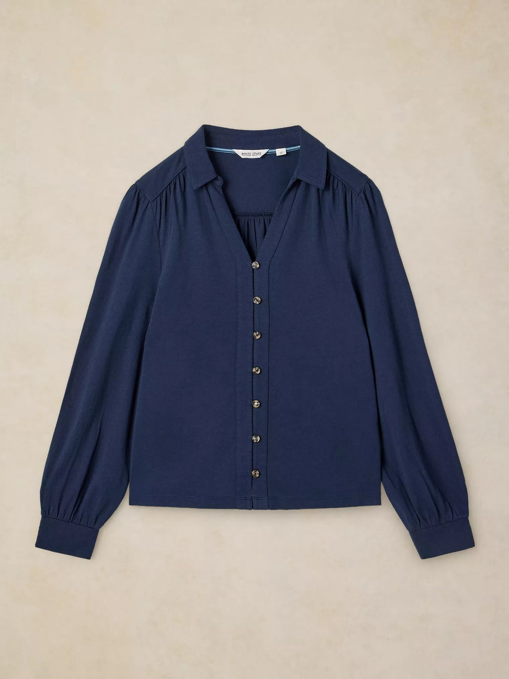 A navy blue long-sleeve blouse with a button-down front, collar, and gathered shoulders, laid flat on a textured beige background.