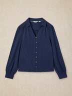 A navy blue long-sleeve blouse with a button-down front, collar, and gathered shoulders, laid flat on a textured beige background.