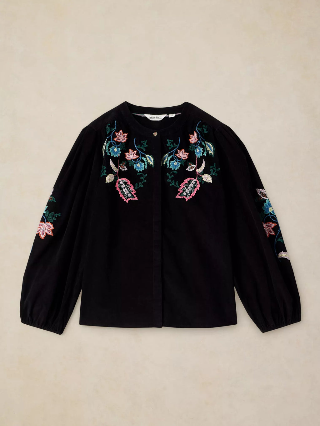 A black long-sleeve button-up blouse with colorful floral embroidery on the chest and sleeves, laid flat on a textured beige background.