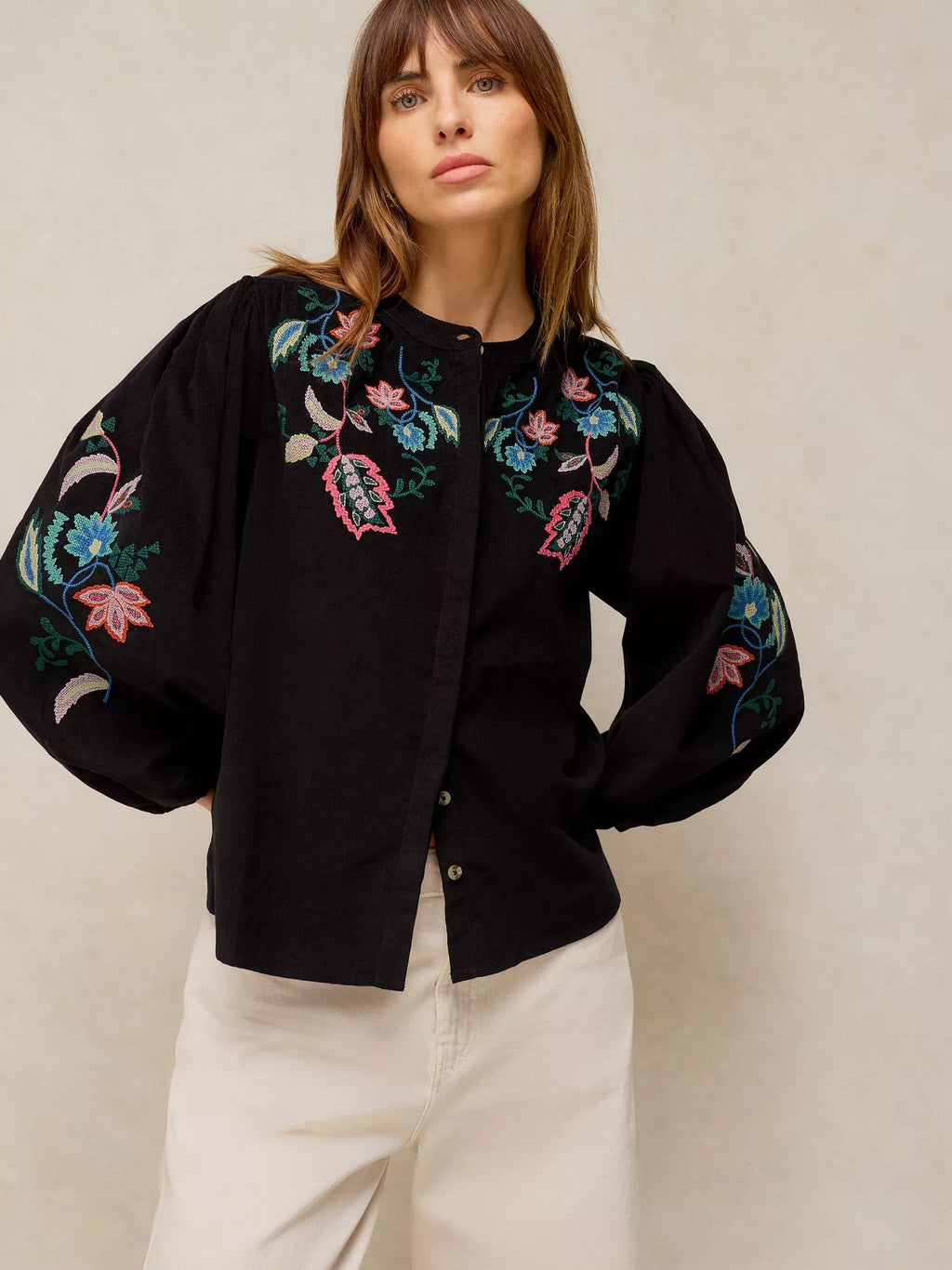A woman wearing a black button-up blouse with colorful floral embroidery on the chest and sleeves, paired with cream-colored trousers, set against a neutral textured beige background.