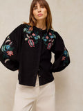 A woman wearing a black button-up blouse with colorful floral embroidery on the chest and sleeves, paired with cream-colored trousers, set against a neutral textured beige background.