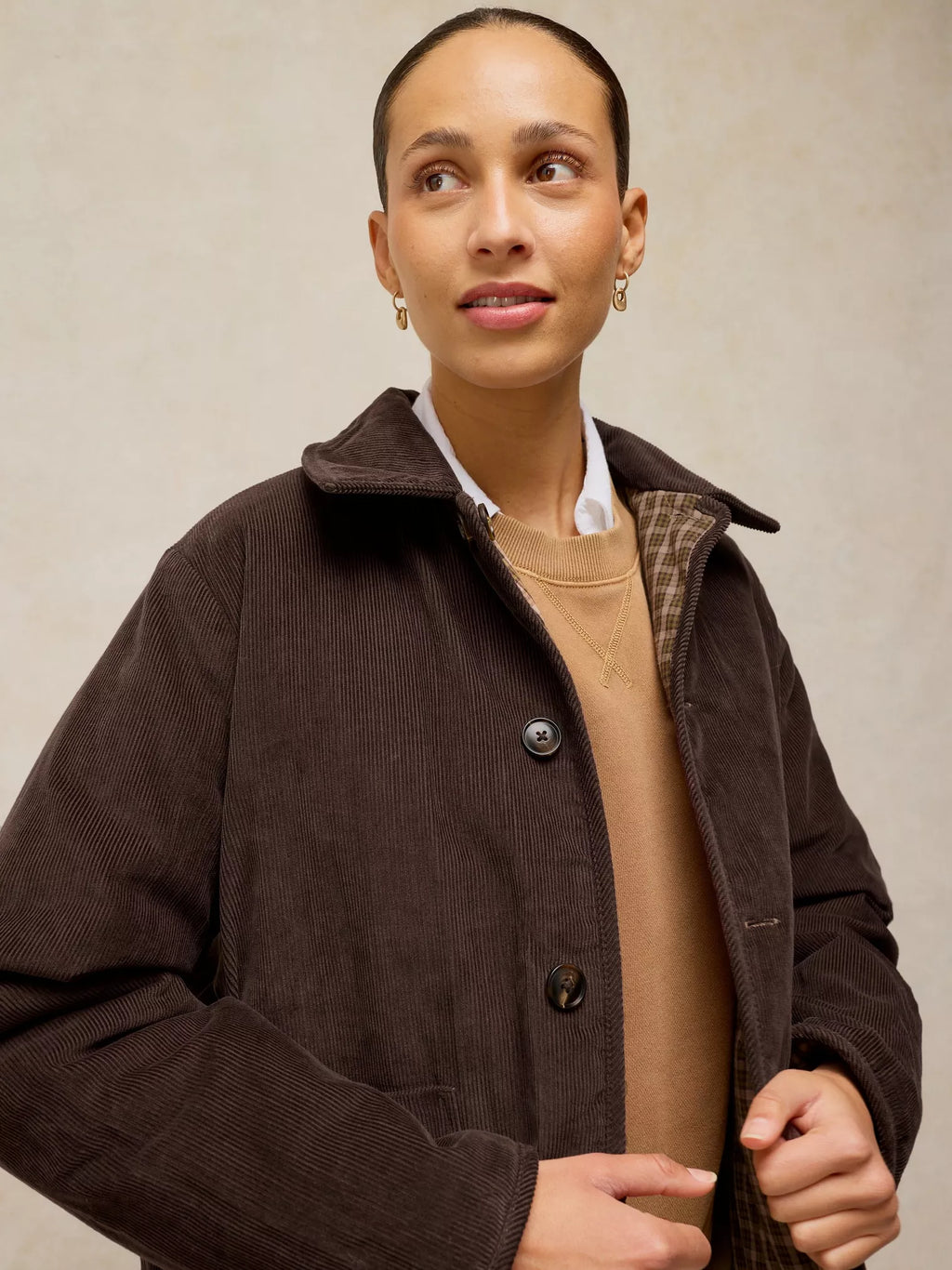 A woman wearing a dark brown corduroy jacket over a beige sweater and white collared shirt, photographed against a neutral beige background.