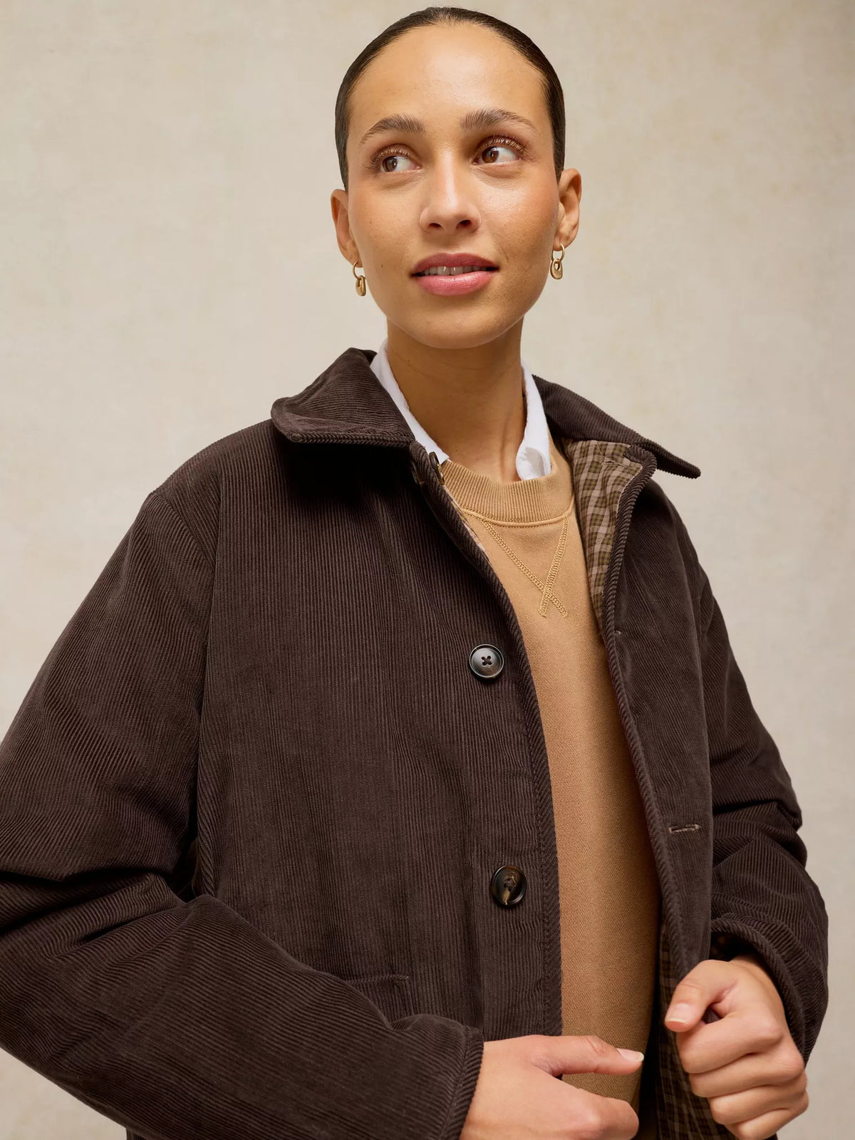 A woman wearing a dark brown corduroy jacket over a beige sweater and white collared shirt, photographed against a neutral beige background.
