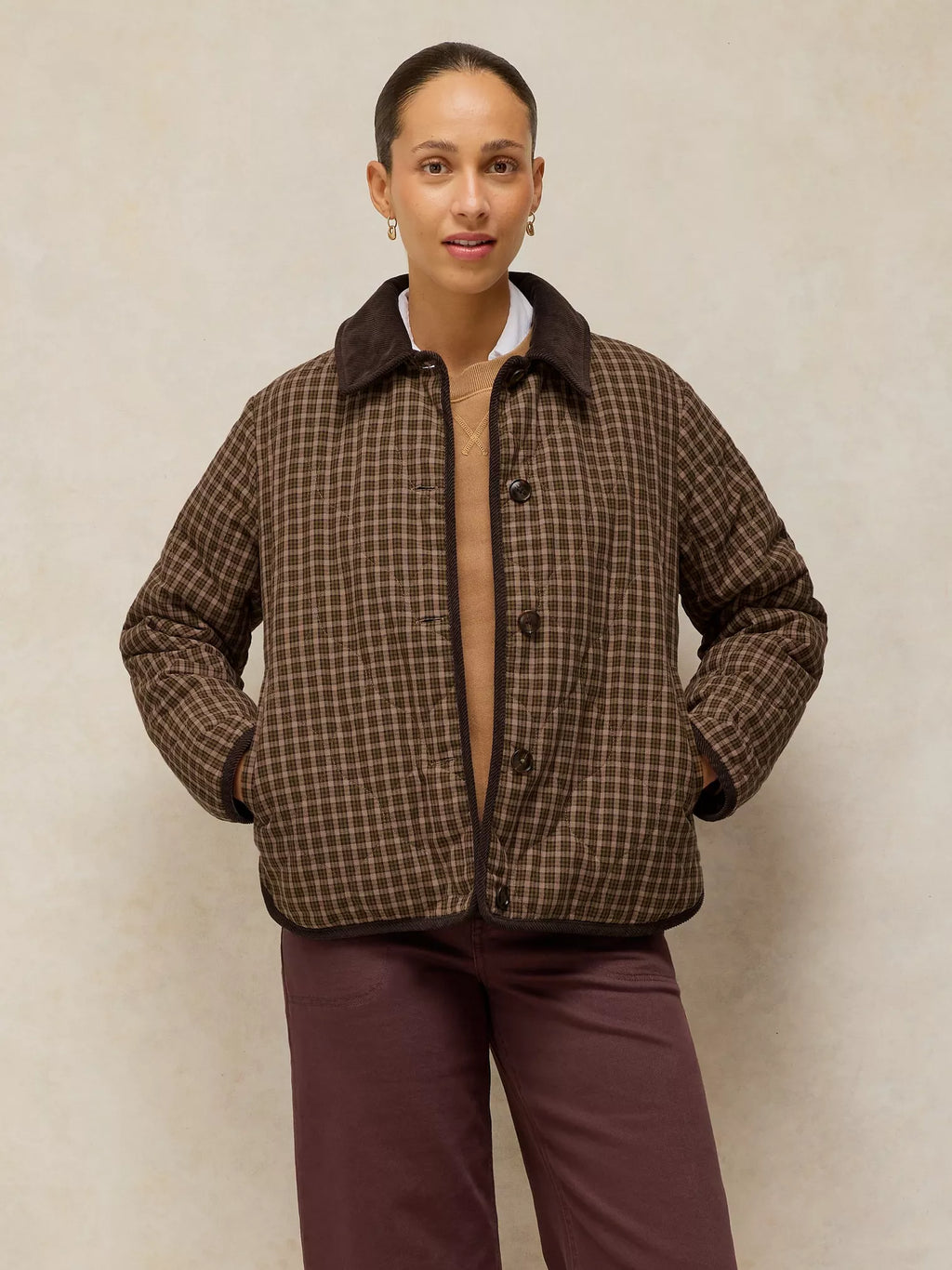 A model wearing a brown and beige plaid quilted jacket with a dark brown corduroy collar, paired with dark plum pants, standing against a textured beige background.