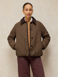 A model wearing a brown and beige plaid quilted jacket with a dark brown corduroy collar, paired with dark plum pants, standing against a textured beige background.